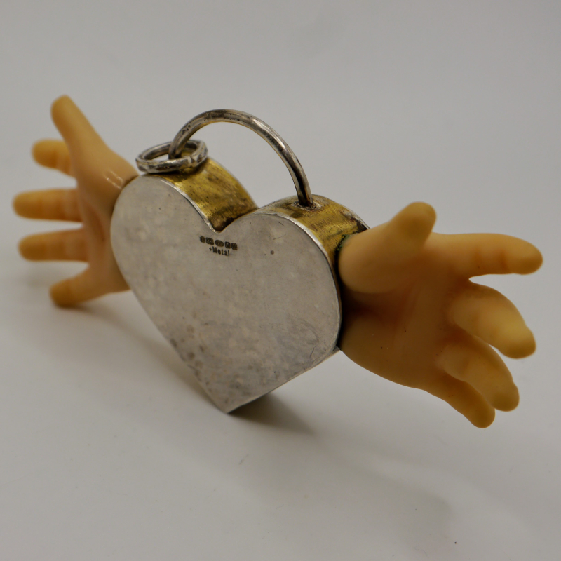 A large silver hollow heart with brass sides and two dolls hand protruding from the sides like wings