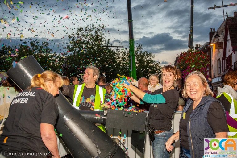 The image shows a group of 3 women and 1 man in a market place loading confetti into a huge confetti blower. 