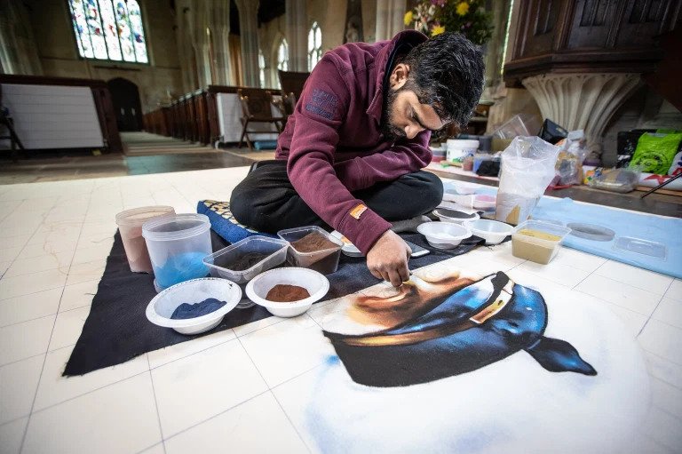 The image shows a young Indian man leaning over a huge picture on the floor of a church. He is creating the picture by sprinkling powder on the floor, this is known as Rangoli 