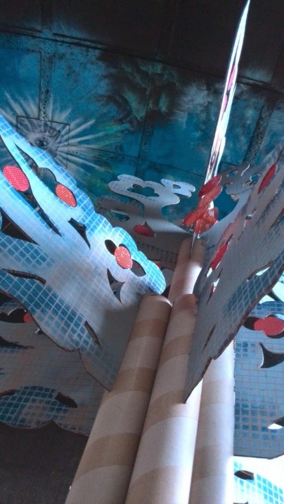 The image shows a view looking up into the canopy of a tree that is made of cardboard. The viewer can see the roof which is painter blue with clouds. 