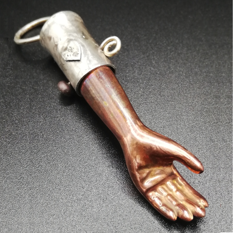 A copper plated dolls forearm is made into a pendant with a silver bezel which has a small silver heart on it.