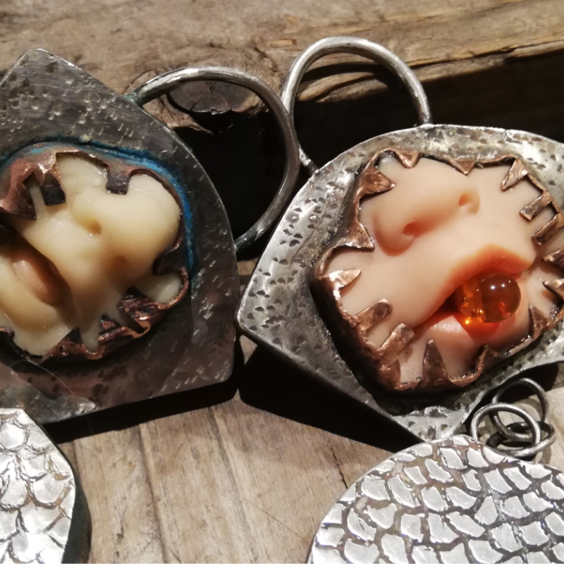 Two dolls faces protrude from heavily textured silver pendants, one has a piece of amber in its mouth