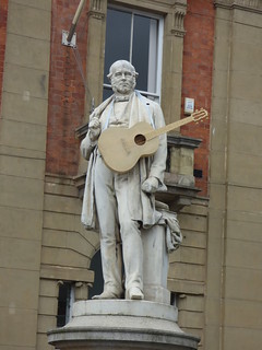 The image shows a statue of Roland Hill with a guitar strung around his neck
